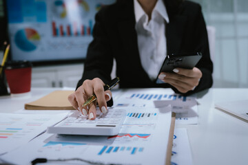 A professional businesswoman calculates financial data using a calculator, surrounded by charts and...