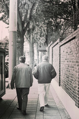 Black and white photo of two elderly men walking along a quiet sidewalk next to a brick wall