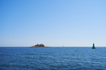 Distant rocky island and green buoy floating on calm blue sea under clear sunny sky