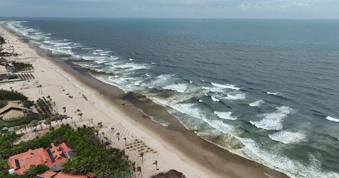 Aerial shot of continuous waves approaching the shore of beach with tourists in Fortaleza, Brazil.