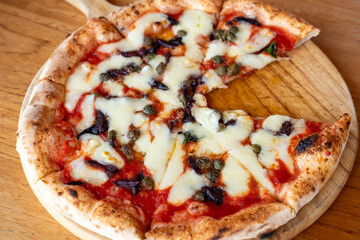 Top view of delicious cheesy pizza on wooden serving board with tomato sauce, olives, capers, and herbs. Perfect for Italian cuisine lovers and food photography.