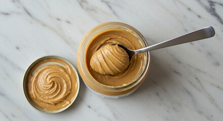Creamy peanut butter in glass jar, spoon, and lid on white marble surface, top view