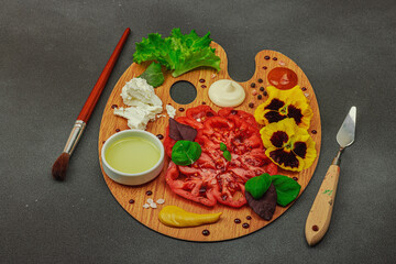 Sliced heirloom tomato with feta, herbs, edible flowers, and sauces on a wooden artist palette