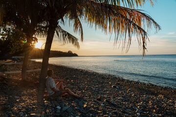 Man relaxing under a palm tree at sunset on a rocky beach in Bali. Peaceful tropical coastline, golden light, and calm ocean create a tranquil summer scene.