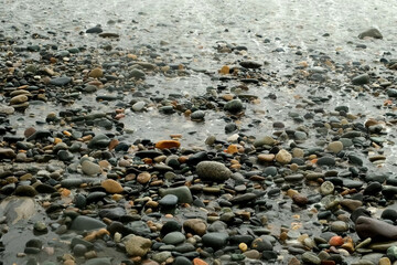 Streaming water from wave on pebble sea beach with white foam, close-up view. Stormy waves washing stone coast in overcast weather. Power of nature and water.