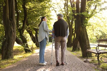 Smiling caregiver supporting elderly man who using walking frame outdoors, back view