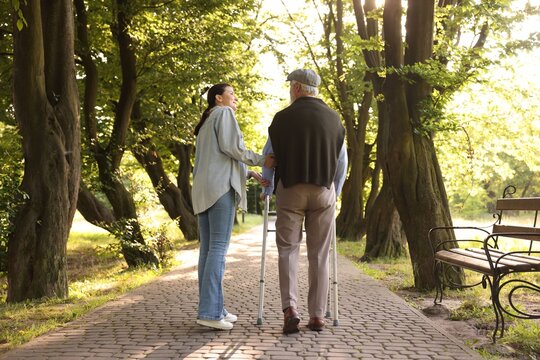 Smiling caregiver supporting elderly man who using walking frame outdoors, back view
