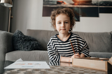 A focused child, with curly brown hair, kneels on the floor, drawing at a glass coffee table in a...