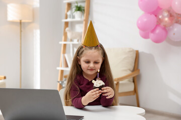 Birthday celebration. Little girl with cupcake having video call via laptop at table indoors