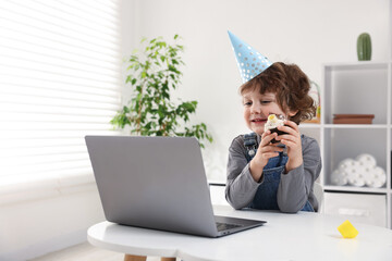 Birthday celebration. Little boy with cupcake having video call via laptop at table indoors. Space...