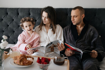 
A joyful child, with curlers in her hair, shares a cozy morning in bed with adults, as they read...