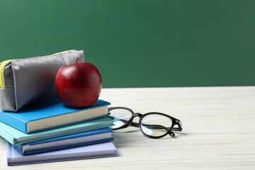 Different notebooks, pencil case, glasses and red apple on white wooden table against blackboard,...