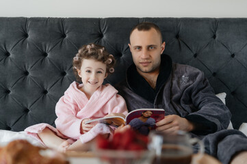 A happy child with curlers in her hair, wearing a pink bathrobe, smiles lovingly at her father in a...