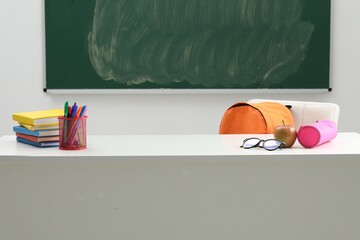 Different school stationery, glasses and apple on white table near blackboard indoors