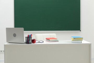 Different school stationery, laptop, glasses and red apple on white table near blackboard indoors