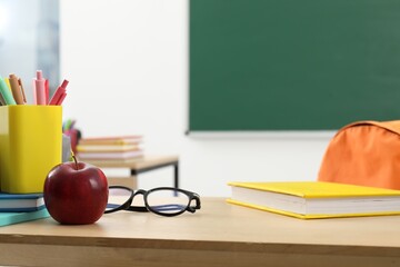 Different school stationery, glasses and red apple on wooden table near blackboard indoors