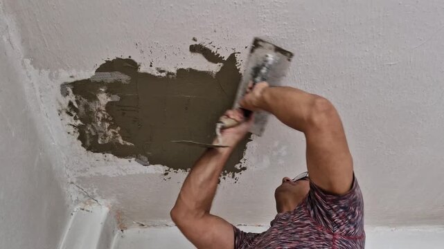 An experienced elderly man carries out a ceiling repair in a residential room by applying adhesive and plaster over reinforcing mesh (perlinka) using hand tools.