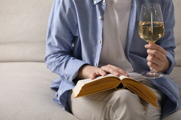 Woman with glass of wine reading book on sofa at home, closeup