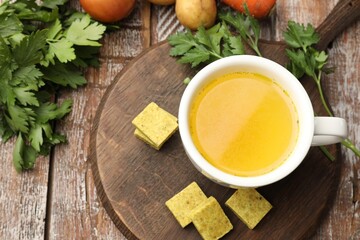 Aromatic bouillon cubes, broth, parsley and vegetables on wooden table, flat lay