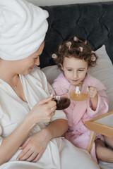  happy child with curlers in her hair, wearing a pink bathrobe, clinks glasses with a woman in a...