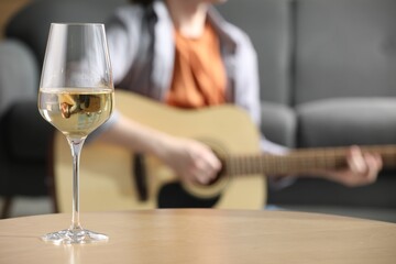 Glass with white wine on table and man playing guitar indoors, selective focus