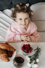 
A child with curlers in her hair, wearing a pink bathrobe, smiles cheerfully while enjoying breakfast in bed with a croissant and fresh raspberries