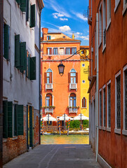 Venice, Italy. View from the narrow street with brick walls of houses at vibrant colorful houses above water of channel. Sunny landscape. Summer day.