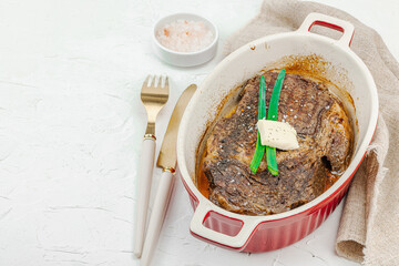 Raw ribeye steak in ceramic baking dish with herbs, spices, cutlery and napkin on white plaster