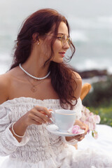 Elegant Woman Enjoying Tea Outdoors in White Dress with Ocean View
