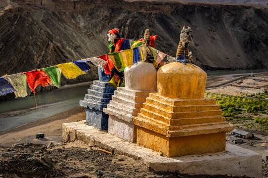 View of prayer flags fluttering above the colorful stupas, contrasting against the arid mountains and winding river, Leh, Ladakh, India.