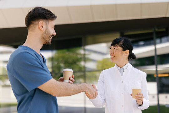 Healthcare professionals shaking hands and smiling while holding coffee cups outdoors at a modern hospital