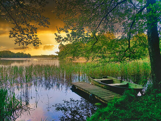 Wooden dock with old rowboat on calm lake surrounded by reeds and trees at golden sunset. Warm sky...