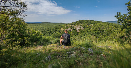 A hiker sitting in the grass, facing the forested cliffs of Nagy-Farkaskő on a warm summer day, surrounded by lush greenery and peaceful views.