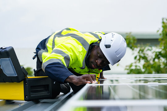 African engineer in safety helmet and reflective jacket installing solar panel on rooftop, focusing on renewable energy technology and precision work
