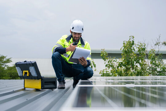 Engineer on rooftop wearing safety vest and helmet checking solar panel system using clipboard and walkie-talkie to log maintenance report during scheduled inspection in sunny weather. - Powered by Adobe