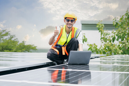 Smiling engineer wearing helmet and safety vest giving thumbs up while working with laptop on rooftop solar panels, symbolizing renewable energy success, clean technology, and sustainable future. - Powered by Adobe