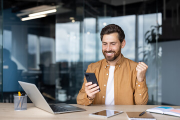 A happy man celebrates his success while looking at his smartphone in the office, a laptop, tablet, and notebook are on the desk.