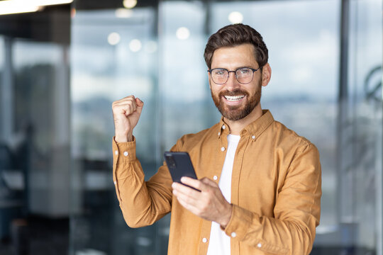 A happy man with a beard looks at his phone with a fist raised in victory.