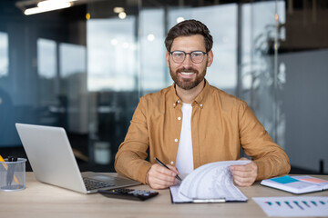 A smiling businessman wearing glasses sits at his desk, reviewing documents while working on his...