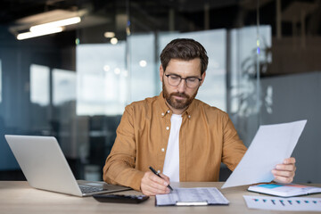 A businessman at his desk reviewing documents while working on a laptop, surrounded by charts,...