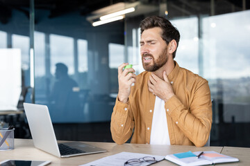 A man in an office uses a throat spray, potentially for a sore throat or cold, near a laptop.