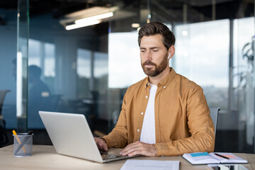 A focused man works on a laptop at a desk in a modern office environment, wearing earbuds.