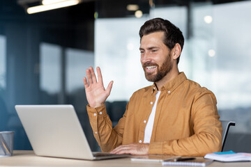 A smiling businessman waves during a video call, sitting in a modern office environment, using a...