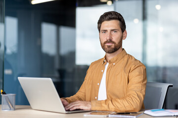 A focused businessman works at a laptop in an office setting. He is looking directly at the camera.