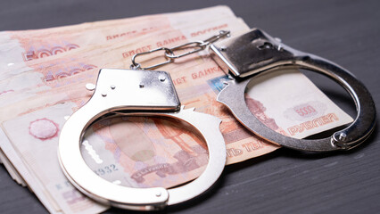 A pair of handcuffs is on top of a stack of money. The handcuffs are silver and appear to be brand...