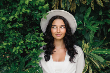 Elegant Brunette Woman in White Hat Posing Among Lush Greenery