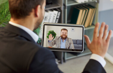 Man in office holding tablet displaying video call smiling, waving, online interaction, virtual...