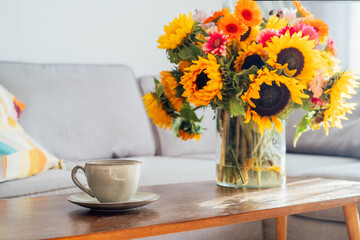 Focus on cup of hot drink and vase with huge sunflowers bouquet on wooden coffee table in modern cozy light living room with gray couch sofa. Cozy Design home decor. Summer mood, Relaxing rituals.