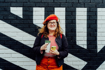 smiling plus size woman with reusable coffee cup and phone on painted wall background. Body positive, self-acceptance. Urban street fashion style. Confident happy satisfied overweight model posing