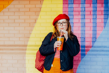 Smiling plus size woman in glasses and bright clothes drinking sugar flavored tapioca bubble tea near urban colorful painted brick wall. Happy fashionable hipster overweight girl. Street fashion
